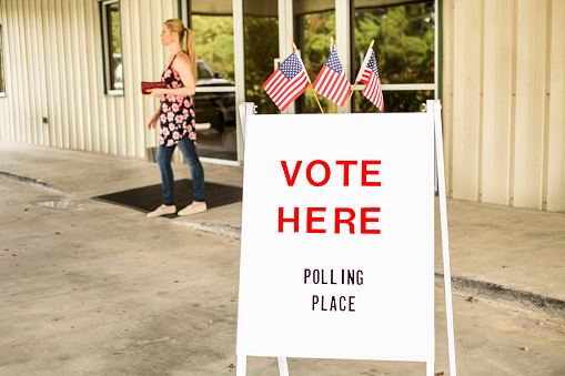 'Vote Here, Polling Place' sign outside of a local, public polling location in USA.  American flags top the sign.  One woman exits the building.  The USA elections are held in November each year.