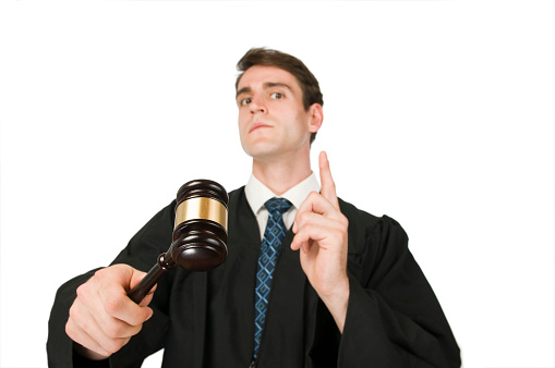 Torso view under view of a young man with black robe and gavel against white background with raised index finger is seriously looking into the camera.
