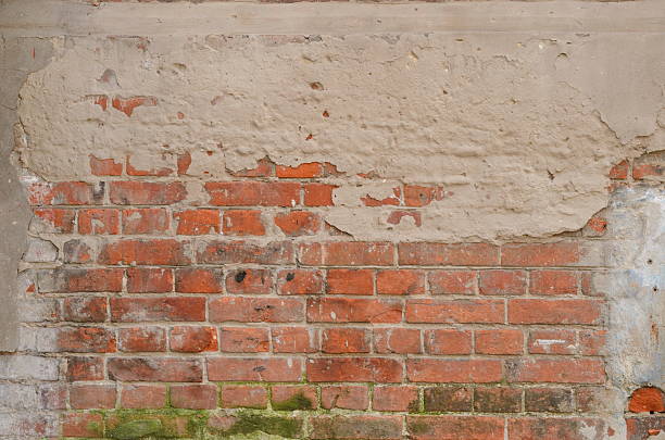 A close-up of an old, abandoned building facade with peeling paint, exposed bricks, and two weathered wooden windows. The cracked plaster and decayed surface highlight the structure's age and deterioration.