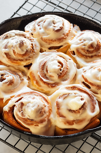 Homemade cinnamon rolls or buns baked in a cast iron skillet and covered with cream cheese icing. The skillet is cooling atop a black wire cooling rack on a white and gray marble countertop.