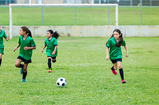 A group of elementary age soccer players run to kick the soccer ball on the playing field. A soccer goal is in the background. They are wearing green uniforms.