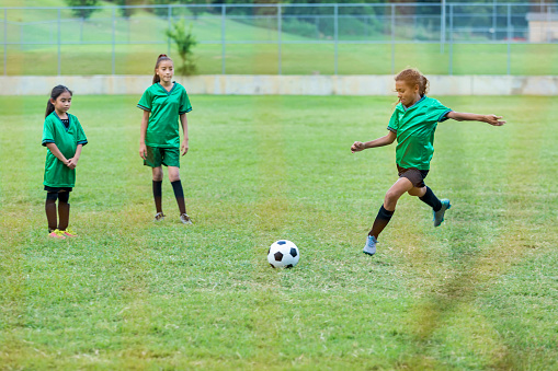 Female athlete prepares to kick soccer ball during her game. She is wearing a green uniform. Her teammates are on the playing field with her.