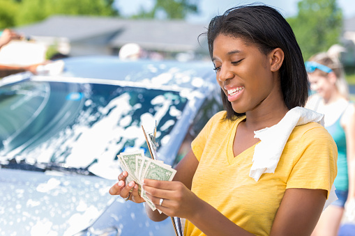 Pretty African American girl smiles while counting the profits from fundraising car wash. She is fanning out the money while counting it. She has a towel on her shoulder. Friends are washing a car in the background.