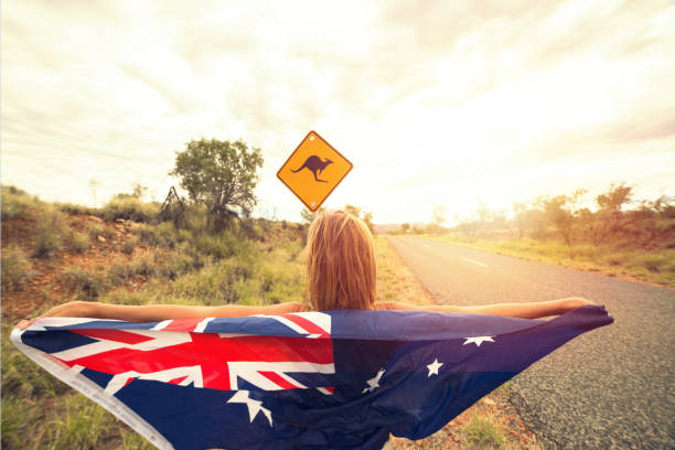 Female in Australia holding flag near kangaroo sign Cheerful young woman on the road standing next to a kangaroo warning sign, Australia. She is holding an Australian flag in the air. kangaroo crossing sign stock pictures, royalty-free photos & images