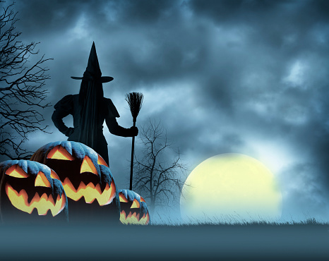 A rear view of a Halloween witch holding her broom as she stands behind a group of jack o' lanterns while she watches a full moon rising over the horizon.