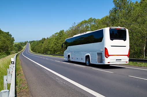 White Bus driving along an empty asphalt road lined with deciduous trees in the countryside