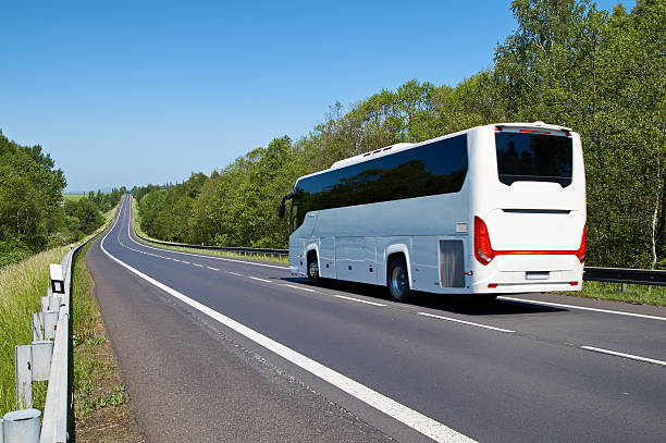 White Bus driving along an empty asphalt road lined White Bus driving along an empty asphalt road lined with deciduous trees in the countryside coach bus photos stock pictures, royalty-free photos & images