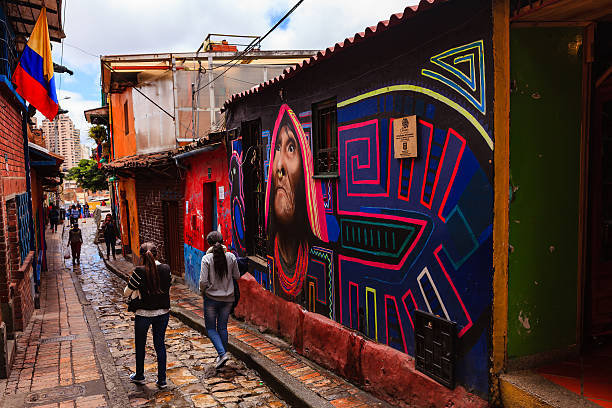 Bogotá, Colombia - Local Colombians Walk Through The Narrow, Colorful, Cobblestoned Calle del Embudo In The Historic La Candelaria District Bogota, Colombia - July 20, 2016: A couple of young Colombian girls walk down the narrow, cobblestoned, Carrera Segunda, in the historic district of La Candelaria in the Old Town of the Andean capital city of Bogota, Colombia, in South America. The City was founded in the 16th Century in this area, by the Spanish Conquistador, Gonzalo Jiménez de Quesada. Many walls in this area are painted with either street art, or legends of the pre Colombian era, in the vibrant colours of Latin America. The wall of a shop on the right is an example of such murals. It has rained a little and the Carrera is wet; the overcast sky promises more rain. 20th July is National Day in Colombia and the country's flag is flown on many buildings. Photo shot in the morning sunlight, on a cloudy day. Horizontal format. calle del embudo stock pictures, royalty-free photos & images