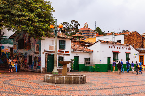 Bogota, Colombia - July 20, 2016: The small square called Chorro de Quevedo in the La Candelaria district of Bogotá, the capital city of the South American country of Colombia. It is here that the Spanish Conquistador, Gonzalo Jiménez de Quesada founded the city in 1538. Many walls in this area are painted with either street art, or legends of the pre Colombian era, in the vibrant colours of Latin America. Some old colonial buildings can be seen on the square. It is from this place that the Zipa, or Chief of the Muisca tribe, viewed the Savannah de Bogota regularly. In 1832 the site was purchased by the Augustinian priest, Father Quevedo, who installed the public water fountain seen in the centre of the square. Its water supply was cut off  when a nearby building collapsed in 1896. Considered a must-visit-site in the city, some tourists and local Colombians can be seen in the image. Photo shot on a cloudy morning; horizontal format. Copy space.