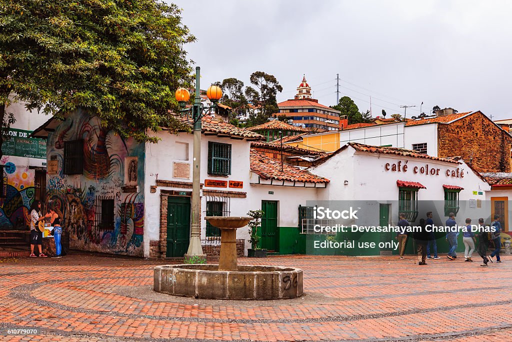 Bogota, Colombia - Tourists on Plaza Chorro de Quevedo Bogota, Colombia - July 20, 2016: The small square called Chorro de Quevedo in the La Candelaria district of Bogotá, the capital city of the South American country of Colombia. It is here that the Spanish Conquistador, Gonzalo Jiménez de Quesada founded the city in 1538. Many walls in this area are painted with either street art, or legends of the pre Colombian era, in the vibrant colours of Latin America. Some old colonial buildings can be seen on the square. It is from this place that the Zipa, or Chief of the Muisca tribe, viewed the Savannah de Bogota regularly. In 1832 the site was purchased by the Augustinian priest, Father Quevedo, who installed the public water fountain seen in the centre of the square. Its water supply was cut off  when a nearby building collapsed in 1896. Considered a must-visit-site in the city, some tourists and local Colombians can be seen in the image. Photo shot on a cloudy morning; horizontal format. Copy space. Bogota Stock Photo