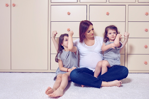 lifestyle shot of pregnant mother near her little daughters playing together and showing, stretching muscles.