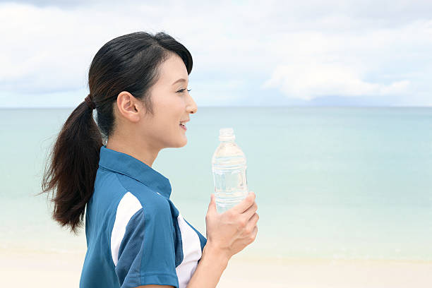 A close-up of a water bottle at the beach, with sunlight reflecting off its surface. The image evokes summer, freshness, and the importance of staying hydrated by the sea
