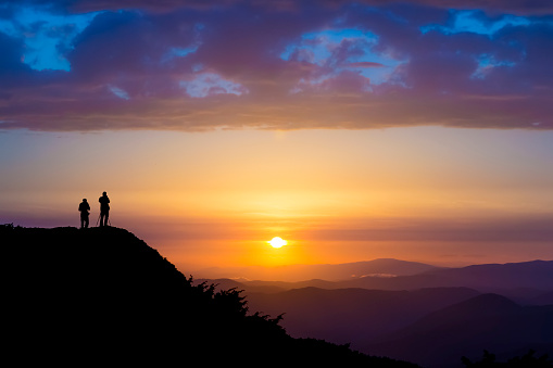 Silhouettes of two people standing on a rock and looking toward the sun. Dramatic clouds over them. Sunset in the mountains. Purple light. Summer in the Ukrainian Carpathians
