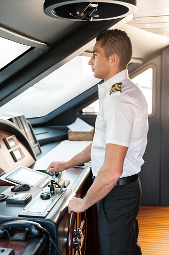 Vertical color image of young boat captain operate a yacht steering wheel in ship's bridge.