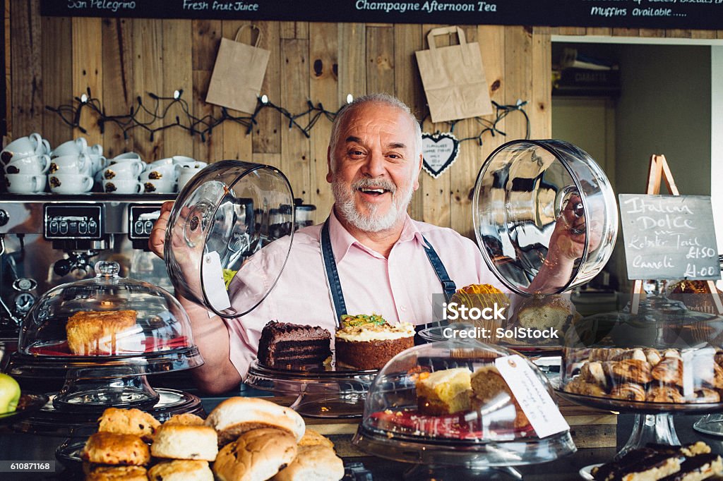 What can I get you? A senior man is standing behind a counter in a coffee shop ready to take an order form a customer. He has a friendly face and is wearing an apron. Cake Stock Photo