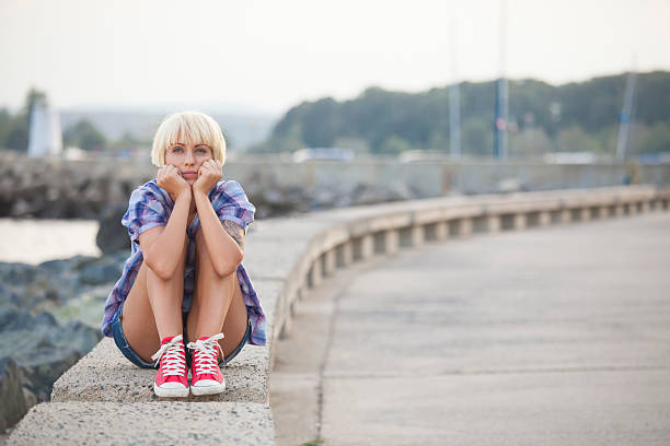 Worried blond woman sitting on parapet with head in hands next to the sea. Hands leaning on knees, wears casual clothes and red sneakers. On background the sea and clif beach. Focus on foreground.