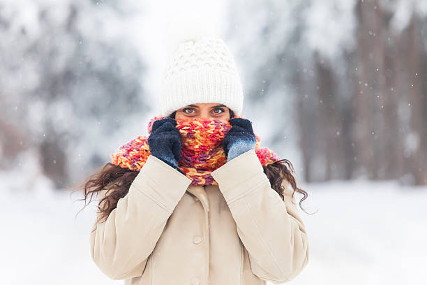Smiling young woman freezing in the snow forest. With winter coat, knit scarf and hat. Hiding face behind her scarf and looking at camera. Snow forest on background. Snowflakes in the air. Focus on foreground.