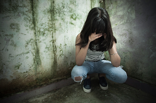 Outdoor image of a serene, sad late teen Asian girl thinking and looking down while leaning against dirty wall. She has long black, straight hair and holding her head in hands. One person, waist up, horizontal composition with copy space and selective focus.