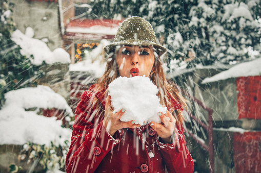 Happy young woman in red winter dress blowing snow below urban scene on cold winter time