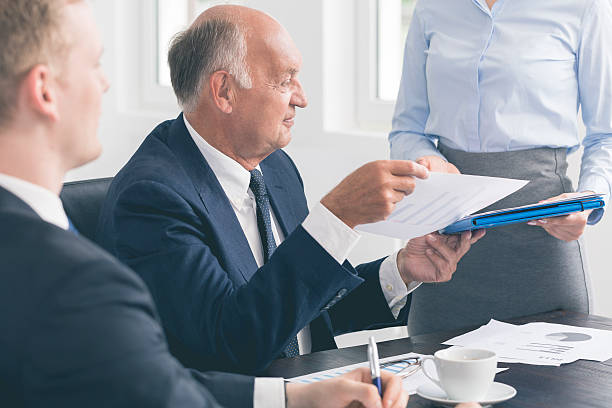 Successful mature businessman assisted by a devoted secretary Close-up of an elegant senior businessman at a desk, with his secretary handing him some documents shareholders meeting stock pictures, royalty-free photos & images