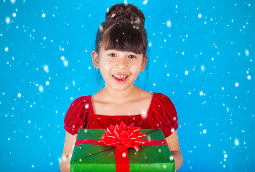 Asian little girl with Santa Claus hat holding big Christmas gift box on blue background.