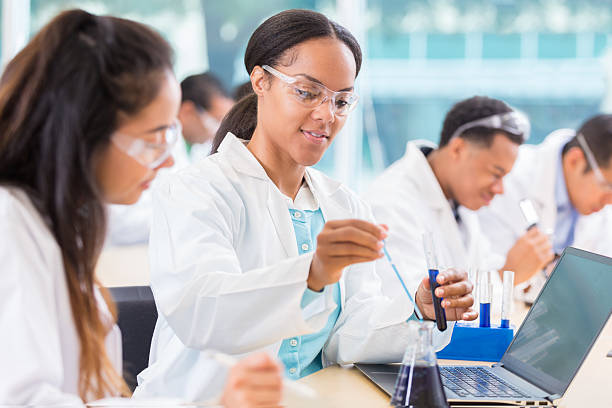 Chemists work on project in lab African American female chemist drops liquid into a beaker. Her assistant is watching her. She is recording the findings on a laptop. beaker photos stock pictures, royalty-free photos & images