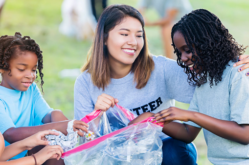 Cheerful Hispanic woman and elementary age African American girls help their neighbors clean up their community. The woman holds a garbage bag while the girls put trash in the bag. The woman has her arm around one of the girls.
