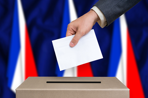 Election in France. The hand of man putting his vote in the ballot box. French flags on background.