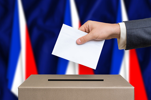 Election in France. The hand of man putting his vote in the ballot box. French flags on background.