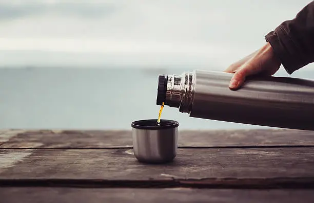 Unrecognizable traveler pouring tea to cup from thermos on wooden table on background of sea. Theme tourism and travel