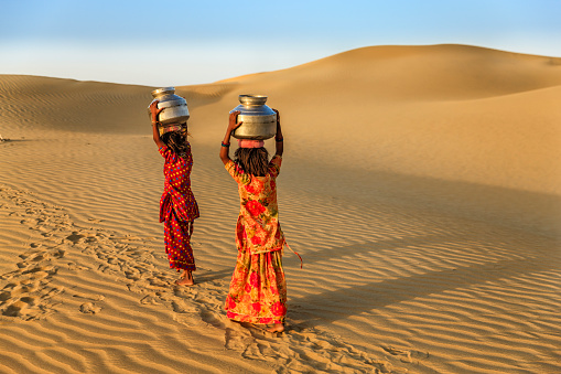 Indian little girls crossing sand dunes and carrying on their heads water from local well, Thar Desert, Rajasthan, India. Rajasthani women and children often walk long distances through the desert to bring back jugs of water that they carry on their heads. 