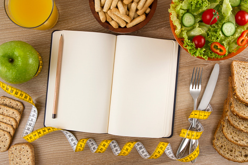 Healthy lifestyle concept with foods on wooden kitchen table with tape measure, and empty notebook.