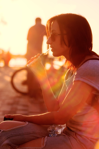 Woman in casual smoking electronic cigarette in sunset light at summer evening. Lifestyle outdoor scene toned in warm color with lens flare and selective focus