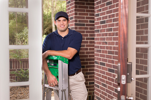 Latin descent repairman or blue collar/service industry worker makes service/house call at customer's front door.  He holds his ladder and wears a blue uniform.  Inspector, exterminator, electrician, plumber.  Red brick home.