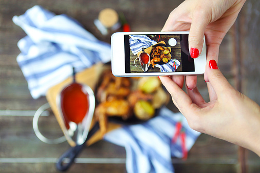 Woman taking a photo of a hot meat dishes on the wooden background with smartphone. Grilled chicken wings with red spicy sauce