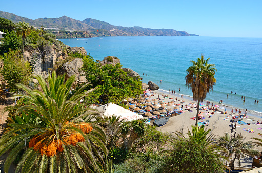 Nerja, Spain - September 18th, 2016: Coastline in Nerja, famous resort in Costa del Sol, called Balcon de Europe. Mediterranean sea on the right. Sierra Nevada mountains on the horizon. In the foreground on right a little beach with straw sunshades and many people. In the foreground on the left rocks ovegrown with many bush and a date palm. Horizontal image in a sunny day.