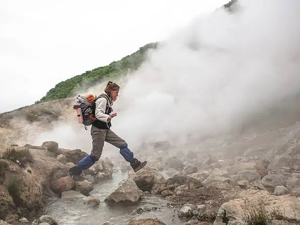 Adult woman with a backpack jumping over a hot stream in a smoking crater of the volcano Mutnovsky on Kamchatka in Russia against the background of a hill with trees and sky with clouds