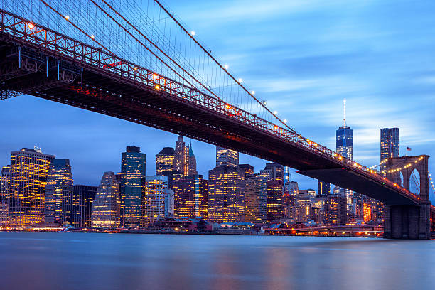  Manhattan skyline and Brooklyn Bridge at dusk. In the foreground is visible East River and Brooklyn Bridge, in the background Lower Manhattan, Manhattan Financial District with illuminated skyscrapers against cloudy, blue sky. On the right side can be seen a new One World Trade Center - Freedom Tower and Beekman Tower the tallest residential tower in the world designed by Frank Gehry. Long exposure with tripod, horizontal orientation, New York City, USA.