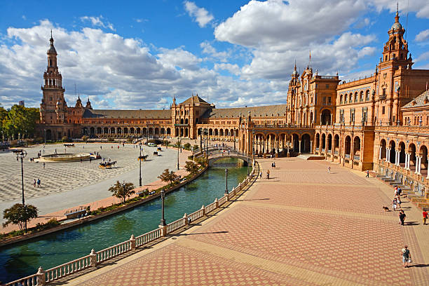 Seville, Spain - September 15th, 2016: Spain Square (Plaza de España) viewed from the southern balcony. Some people walking in the square. The central fountain on the left. Northern tower in the background on the left. Main building on the right. The channel in the middle. Horizontal, wide angle image in a sunny day with dramatic clouds.