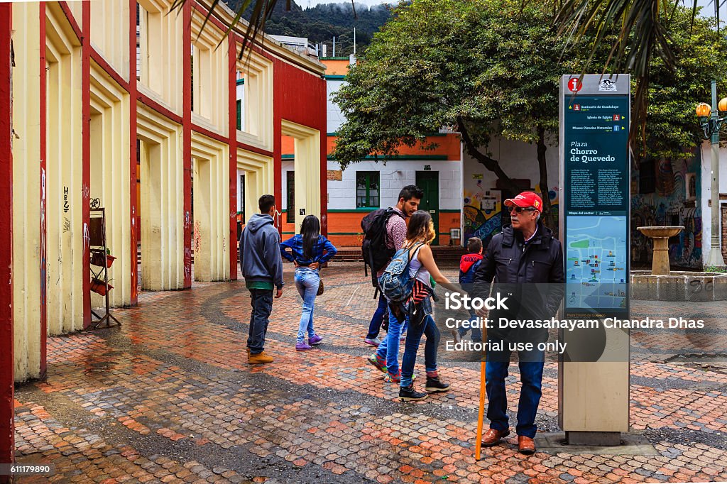 Bogota, Colombia - Tourists on Plaza Chorro de Quevedo Bogota, Colombia - July 20, 2016: The small square called Chorro de Quevedo in the La Candelaria district of Bogotá, the Andean capital city of the South American country of Colombia. It is here that the Spanish Conquistador, Gonzalo Jiménez de Quesada founded the city in 1538. Many walls in this area are painted with either street art, or legends of the pre Colombian era, in the vibrant colours of Latin America. It is from this place that the Zipa, or Chief of the Muisca tribe, viewed the Savannah de Bogota regularly. In 1832 the site was purchased by the Augustinian priest, Father Quevedo, who installed the public water fountain seen to the right of the image. Its water supply was cut off  when a nearby building collapsed in 1896. Considered a must-visit-site in the city, some tourists and local Colombians can be seen in the image. It has rained a little: the square is wet. Photo shot on a cloudy morning; horizontal format.  Andes Stock Photo