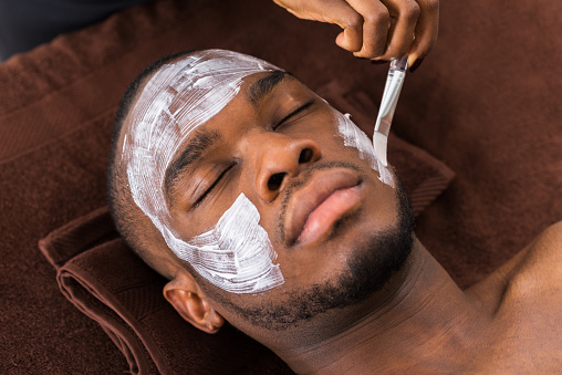 Therapist Applying Face Mask To Young African Man In Spa