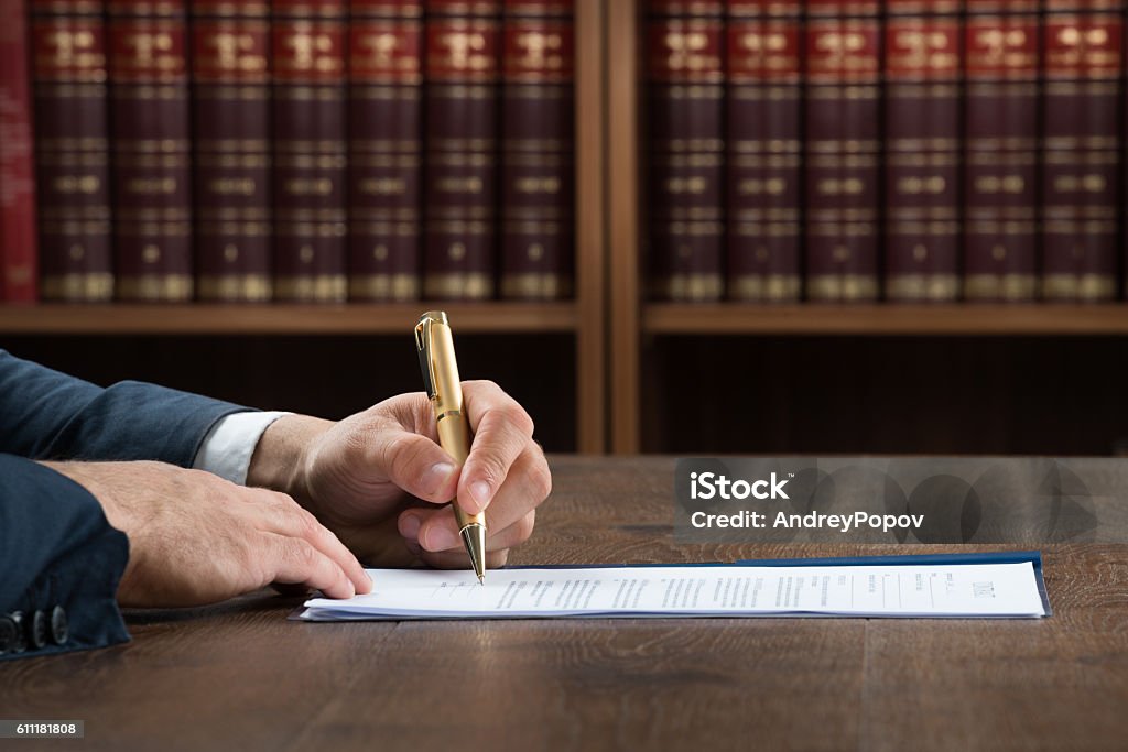 Lawyer Writing On Legal Documents At Desk Cropped image of male lawyer writing on legal documents at desk in courtroom Book Stock Photo