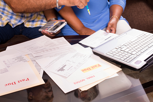 Close-up view of African descent, senior adult couple work together to pay their monthly bills.  They are calculating expenses versus budget income.  Many invoices on living room table.  Great imagery for election season:  home finances, recession, past due bills, mortgage, debt, stress, worry, taxes.