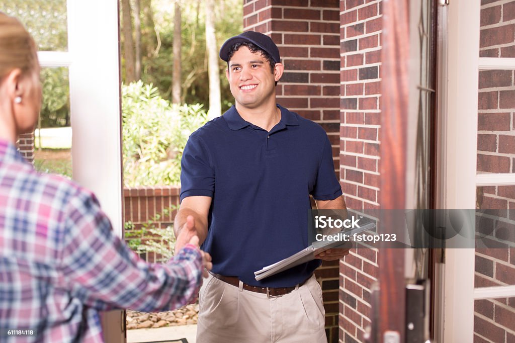 Repairman or delivery person at customer's front door. Latin descent blue collar/service industry worker or delivery person makes service/house call at customer's front door.  He holds a clipboard while shaking hands with customer.  The man wears a blue uniform.  Inspector, exterminator, electrician, plumber, delivery driver.  Red brick home. Residential Building Stock Photo