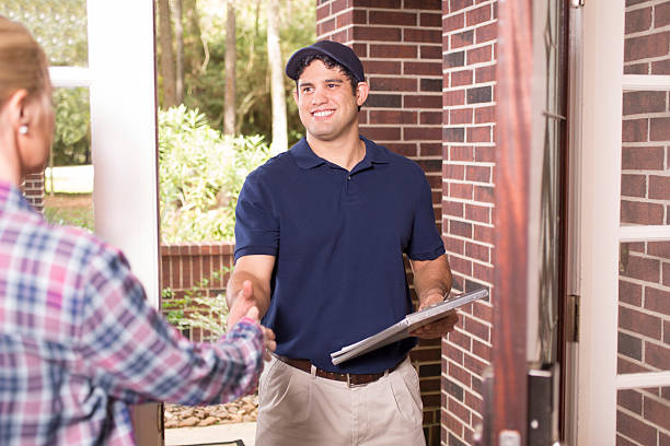 Repairman or delivery person at customer's front door. Latin descent blue collar/service industry worker or delivery person makes service/house call at customer's front door. He holds a clipboard while shaking hands with customer. The man wears a blue uniform. Inspector, exterminator, electrician, plumber, delivery driver. Red brick home. door repairman men manual worker stock pictures, royalty-free photos & images