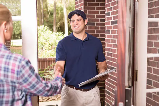 Latin descent blue collar/service industry worker or delivery person makes service/house call at customer's front door.  He holds a clipboard while shaking hands with customer.  The man wears a blue uniform.  Inspector, exterminator, electrician, plumber, delivery driver.  Red brick home.