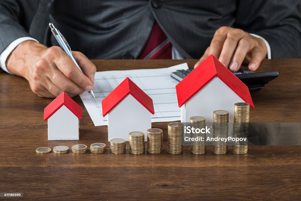 Businessman Calculating Tax By House Models And Coins Midsection of businessman calculating tax by different size houses and stacks of coins on table House Stock Photo