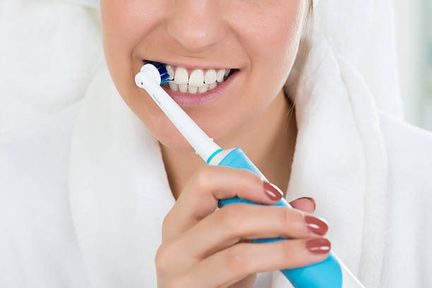 femme en peignoir brossant les dents avec une brosse à dents électrique - human mouth human teeth indoors young women photos et images de collection