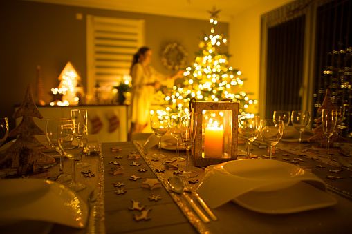focus on table covered for christmas dinner, woman decorating xmas tree in back of living room blurred