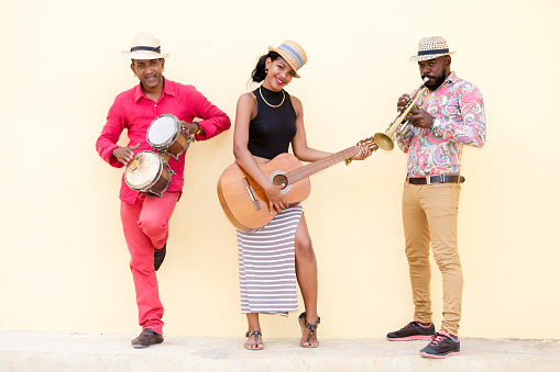 Cuban musical band, the trio consisting of a well known musicians standing against the bright yellow wall. Beautiful young woman standing in the middle, playing guitar. The man on the left holding the small drums bongos, and a musician on the left playing trumpet. Havana, Cuba, 50 megapixel image.